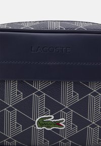 Sötétkék textúrájú táska fehér geometriai mintával. A Lacoste logó és egy zöld, piros hímzett krokodiltövis található rajta.