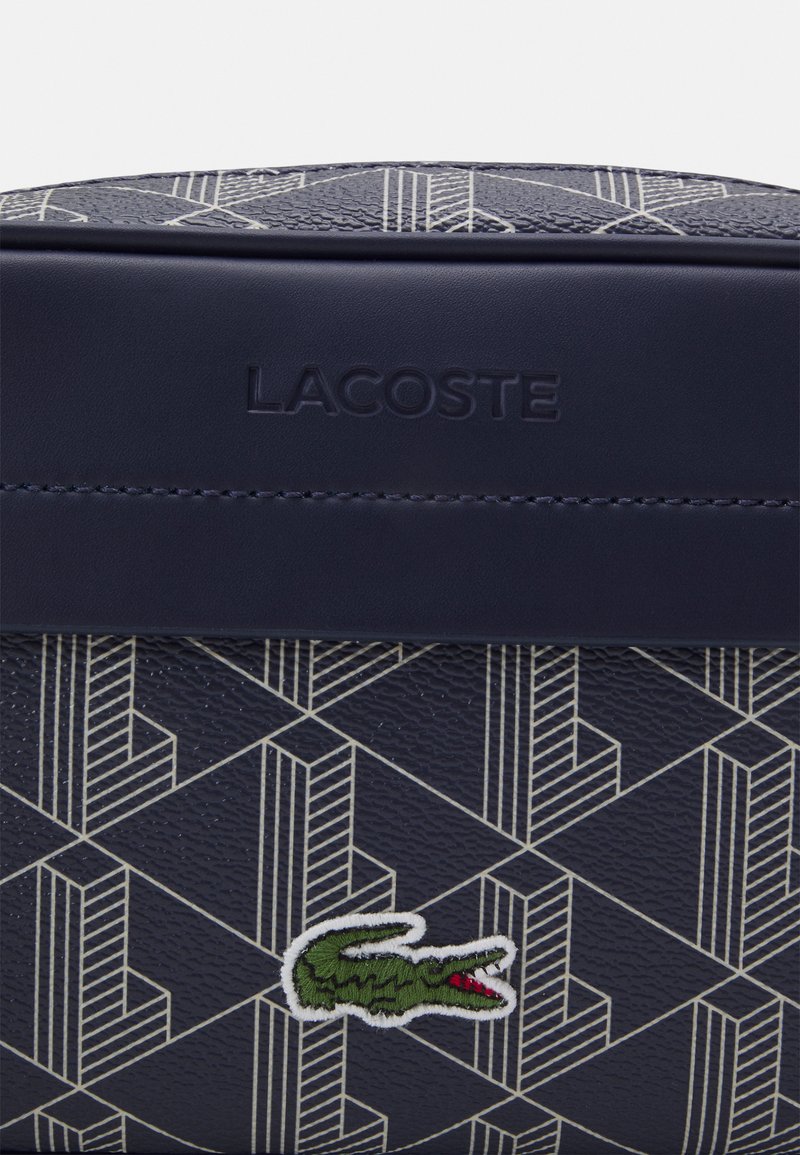 Sac texturé bleu marine avec un motif géométrique blanc. Présente le logo Lacoste et un emblème de crocodile brodé en vert et rouge.