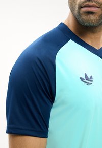 Férfi világoskék és sötétkék rövid ujjú sportpólóban, amelyen egy kis Adidas logó látható a mellkason, válltól állig látható.