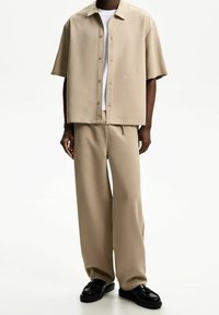 Beżowa koszula z krótkim rękawem o prostym kroju z frontowymi guzikami, w zestawie z dopasowanymi spodniami typu wide-leg. Czarne buty dopełniają całość.