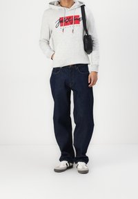 Ljusgrå huvtröja med röd och blå grafik, mörkblå jeans och beige sneakers med svarta detaljer. Liten svart crossbody-väska.