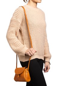 Borsa a tracolla in pelle color cuoio con chiusura a pattina e tracolla regolabile, tenuta contro un maglione beige chiaro con texture e pantaloni neri.