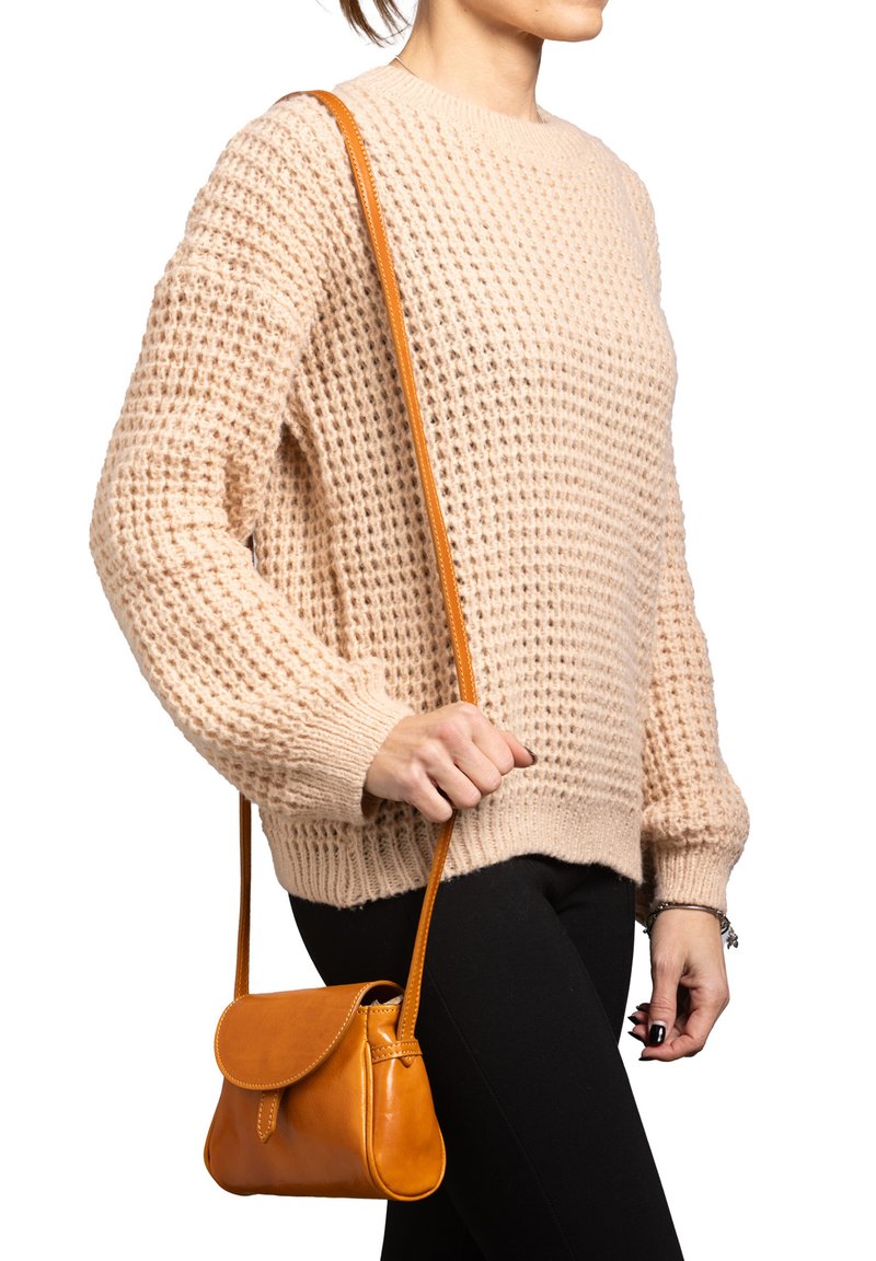 Borsa a tracolla in pelle color cuoio con chiusura a pattina e tracolla regolabile, tenuta contro un maglione beige chiaro con texture e pantaloni neri.