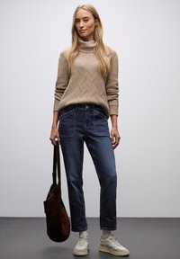 Beige pletený svetr s vysokým límcem, tmavě modré rovné džíny, béžové tenisky a hnědá kabelka přes rameno, umístěné na jednobarevném pozadí.