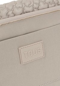 Beżowa torba płócienna z teksturowanym skórzanym wierzchem, z wyróżniającymi się kształtami serc, złotym suwakiem i prostokątną naszywką z logo.