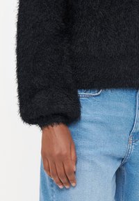 Zwarte fuzzy trui met een losse pasvorm, voorzien van brede manchetten en een zachte textuur, gecombineerd met lichtblauwe jeans. De stof lijkt warm en comfortabel.