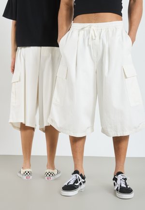 White cargo shorts with a loose fit, featuring side pockets, elastic waistband, and a mid-thigh length. Worn with contrasting sneakers.