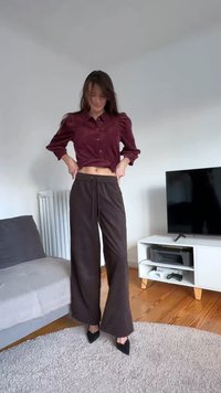 Bordó gombos ing pöttyös ujjakkal, laza szabású sötétbarna nadrággal párosítva. Fekete hegyes sarkú cipő. Semleges háttér egy televízióval.