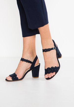 Pieds portant des sandales bleu marine à talon bloc avec des brides festonnées et des boucles, associées à un pantalon court bleu marine sur fond blanc.