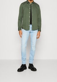 Man i olivgrön knapp-up skjorta över svart topp, ljusblå jeans och svarta snörade stövlar som står mot en neutral bakgrund.