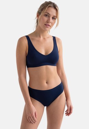 Kvinde stående iført navyblå bh og matchende undertøj foran hvid baggrund, let vendt mod venstre med afslappet udtryk.