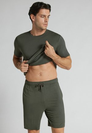 Man tilt een olijfgroene T-shirt op en toont zijn buik, draagt bijpassende shorts met trekkoord, staat tegen een lichtgrijze achtergrond.