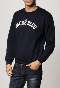 Felpa blu navy in cotone, con collo rotondo e polsini a costine, con la scritta "SACRÉ BLEU" stampata in giallo sul petto.