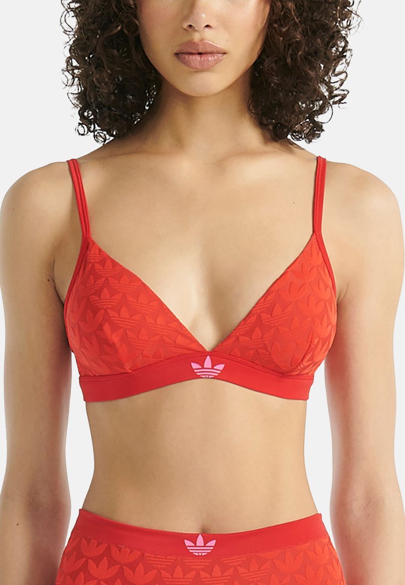 Frau mit lockigem Haar, die einen roten Adidas-Bralette und passende Unterteile mit eingeprägten Trefoil-Logos trägt.