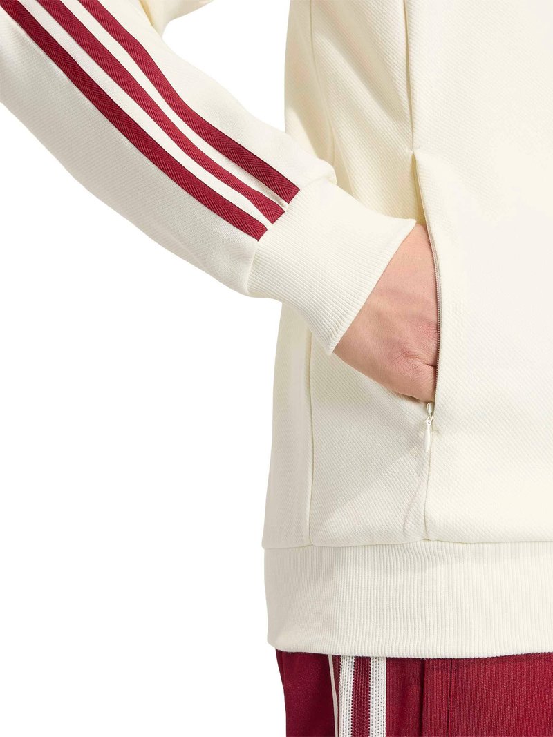 Manche de veste blanche avec trois bandes bordeaux, main dans la poche latérale, associée à un pantalon bordeaux avec des bandes blanches.