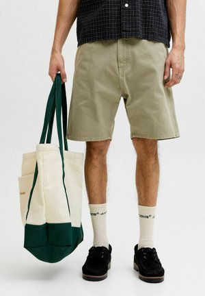 Personne portant un short beige, des chaussures noires, des chaussettes blanches avec du texte, tenant un grand sac fourre-tout blanc et vert par les poignées.