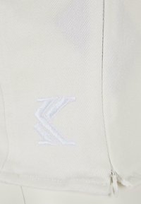 Vit tyg med en subtil ribbad textur, med en vit broderad logotyp vid fåll och en liten dragkedjedetalj på sidan.