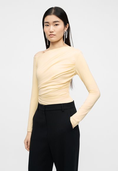 Γυναίκα που φοράει ανοιχτό κίτρινο ασύμμετρο τοπ με ρυτιδώσεις, μαύρο παντελόνι με ψηλή μέση και μακριά κρεμαστά σκουλαρίκια με ρινίσματα, στέκεται με το ένα χέρι στην τσέπη.