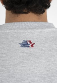 Gros plan sur l'encolure arrière d'un vêtement gris avec un petit motif abstrait brodé en bleu et rouge.
