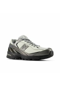 Grå och vita sportskor med mesh-ovandel, vadderad krage och framträdande "N"-logotyp. Texturerad sula för grepp och stabilitet, förstärkt tå.