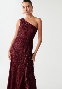 Večerní šaty burgundské barvy se zdobením z flitrů, jednoramenným designem, volánkovými detaily na straně a vysokým rozparkem, které mají přiléhavý střih.