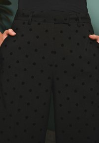 Pantaloni neri con una finitura testurizzata, caratterizzati da pois neri in rilievo. Il design include tasche laterali e una vita con cintura.