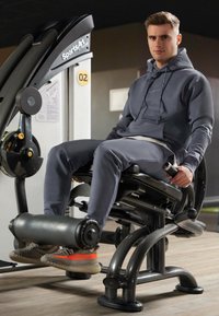 Fitnessapparatuur van SportsArt, met een zwart metalen frame, een verstelbare zitting en een beenpersmechanisme. Gebruiker in grijze kleding gebruikt de machine.