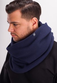 Navy knitted snood with a textured pattern and a folded design. Features a small label with white stitching. Worn over a black shirt.