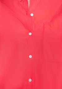 Chemise rouge avec des boutons blancs et une poche poitrine, boutonnée sauf au niveau du bouton supérieur, mettant en valeur la texture du tissu et les détails des coutures.