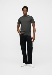 Νεαρός άνδρας που στέκεται φορώντας σκούρο γκρι κοντομάνικο μπλουζάκι με μικρό λογότυπο, μαύρο παντελόνι και λευκά αθλητικά παπούτσια σε απλό λευκό φόντο.