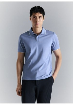 Hombre con camiseta tipo polo de manga corta azul claro y pantalones negros, de pie con una mano en el bolsillo contra un fondo gris liso.