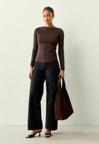 Ruskea pitkähihainen paita, jossa on gathered-yksityiskohta, yhdistetty laajoihin mustiin farkkuihin. Pitelee ruskeaa mokkabagia, jalassa mustat korkokengät.