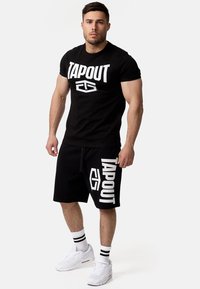 Melna T-krekla un šortu komplekts, ar lielu baltu "TAPOUT" logotipu un grafikām. Baltas sporta kurpes un melnbaltas svītrains zeķes.
