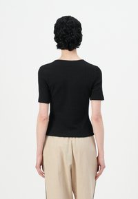Kort zwart geribd gebreid topje met een ronde hals en korte mouwen, gecombineerd met beige broeken, gezien vanuit de achterkant.