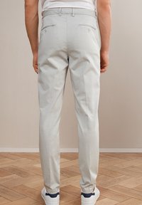 Ljusgrå, skräddarsydda byxor med en slät textur, smal passform och två bakfickor. Kombinerat med vita sneakers och placerat på ett trägolv.