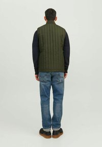Olivengrüne, gesteppte Weste mit hohem Kragen, kombiniert mit einem navyblauen Langarmhemd und blauen Jeans. Schwarze Schuhe mit braunen Sohlen.