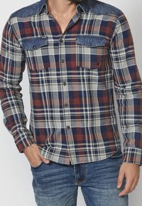 Homme portant une chemise à manches longues à carreaux avec un col en denim bleu et des poches poitrine, associée à un jean bleu, debout avec une main dans la poche.