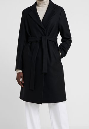 Sort bæltefrakke over beige rullekravesweater, båret med hvide bukser, hænder delvist i lommerne, stående mod en ensfarvet baggrund.