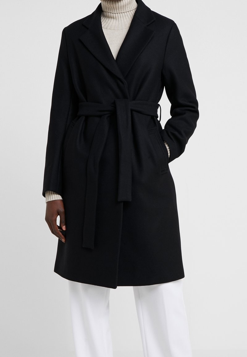 Manteau noir ceinturé sur un pull à col roulé beige, porté avec un pantalon blanc, mains partiellement dans les poches, debout devant un fond uni.