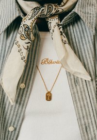 Gestreiftes Hemd mit Knöpfen über einem weißen Rippenoberteil mit "Blkvis"-Stickerei, ein Paisley-Schal um den Hals gebunden und eine goldene Halskette mit Palme-Anhänger.