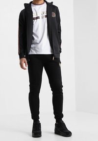 Black zip-up jacket with red and white stripes, white t-shirt with logo, black joggers, and black athletic shoes. Simple, modern design.