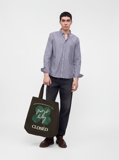 Jeune homme portant une chemise rayée boutonnée et un pantalon foncé, tenant un sac fourre-tout noir avec un trèfle vert et un texte blanc.
