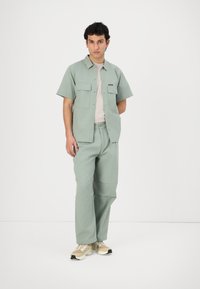 Light green short-sleeve shirt with two chest pockets, paired with matching pants. Pants have a relaxed fit. Model wears beige sneakers.