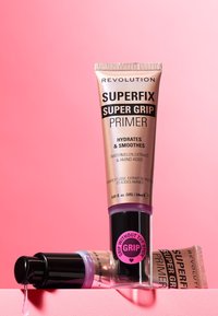 Revolution Superfix Super Grip Primer i en gull- og svart tube, med detaljer av vannmelonekstrakt, på en rosa bakgrunn med en dråpe av produktet synlig.