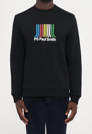 Sweat-shirt noir à manches longues avec des rayures verticales colorées au-dessus du logo "PS Paul Smith" sur la poitrine, porté par une personne debout.