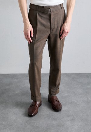 Brown tailored trousers with a textured fabric, featuring pleats and a belt loop. Paired with brown leather loafers with horizontal accents.