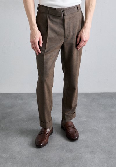 Brown tailored trousers with a textured fabric, featuring pleats and a belt loop. Paired with brown leather loafers with horizontal accents.