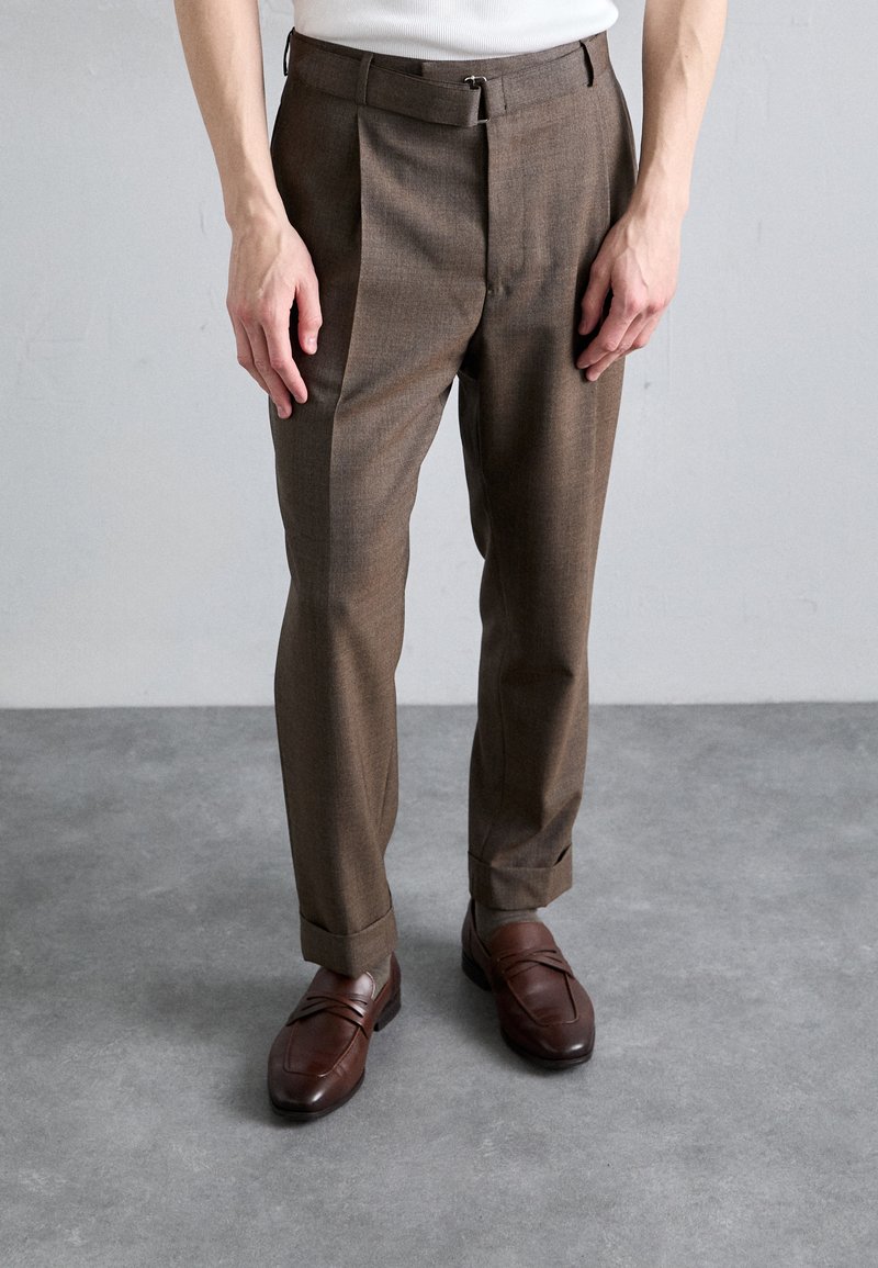 Pantalons marron sur mesure avec un tissu texturé, présentant des plis et des passants de ceinture. Associés à des mocassins en cuir marron avec des accents horizontaux.