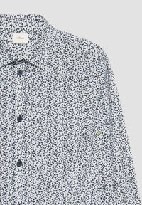 Witte shirt met een klein marinebloem patroon, button-down kraag en marineblauwe knopen. Heeft een klein gele accent nabij de zoom.