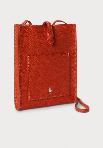 Červená kožená kabelka s texturovaným povrchem, vybavená přední kapsou a ozdobou s logem. Obsahuje malý popruh na držení.